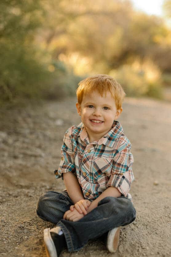 A young boy with red hair sits cross-legged on a dirt path outdoors, smiling at the camera. He wears a plaid shirt and jeans, with greenery and soft sunlight in the background. by Justin Salem Meyer