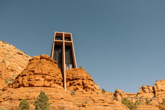 A modern chapel with tall glass windows is built into red rock formations under a clear blue sky, surrounded by rugged desert landscape and scattered green shrubs. by Justin Salem Meyer