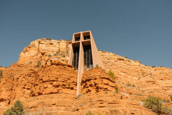A modern church, the Chapel of the Holy Cross, is built into red sandstone cliffs under a clear blue sky, blending with the surrounding rocky landscape. by Justin Salem Meyer