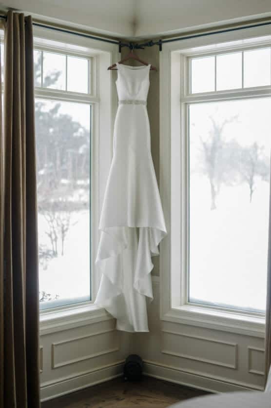 A white sleeveless wedding dress with a fitted bodice and flowing skirt hangs on a wooden hanger in the corner of a room, framed by two tall windows overlooking a snowy landscape. by Justin Salem Meyer