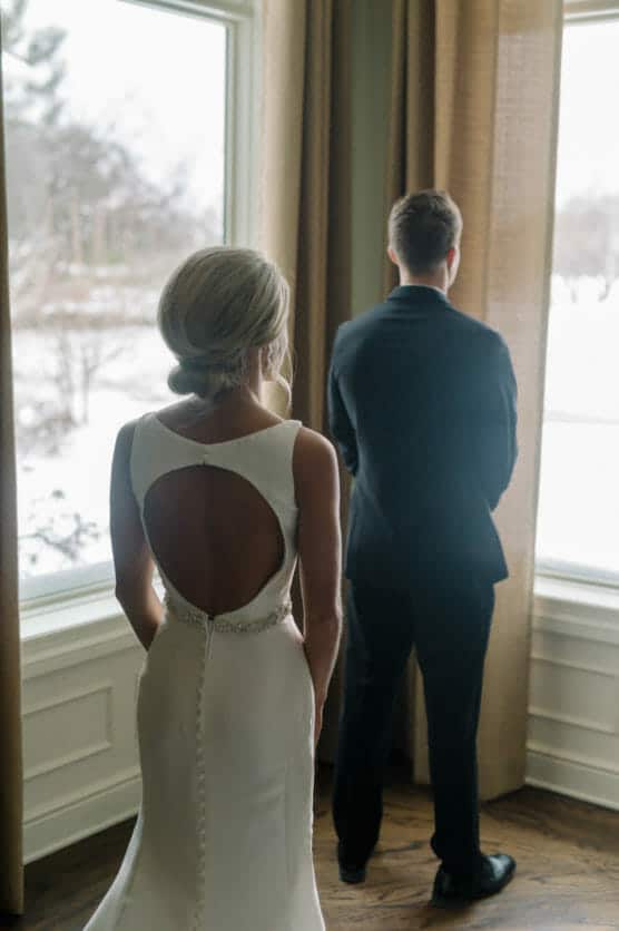 A bride in a white dress with an open back stands behind a groom in a black suit, both facing large windows with a snowy landscape outside. by Justin Salem Meyer