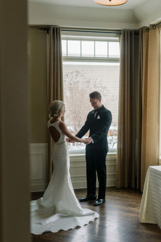 A bride in a white gown and a groom in a black tuxedo stand facing each other, holding hands by a large window with beige curtains, bathed in soft natural light. by Justin Salem Meyer