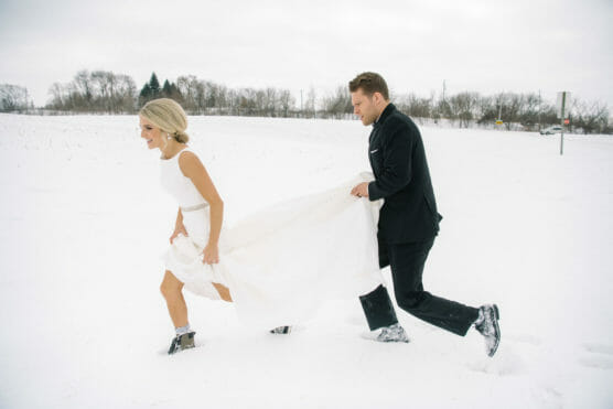 A bride in a white dress and groom in a black suit walk through deep snow; the groom holds up the brides train as they smile and make their way across the wintry landscape. by Justin Salem Meyer