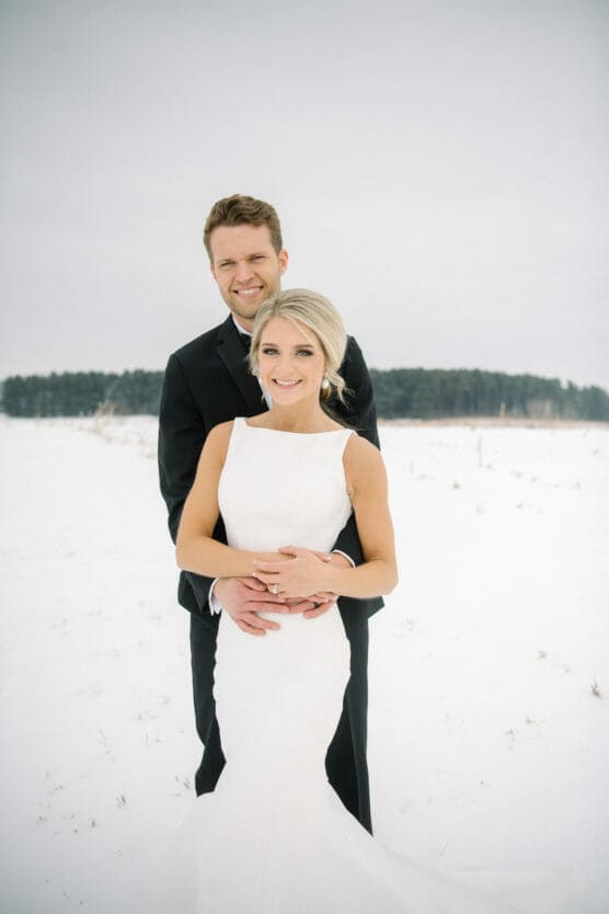 A bride in a white dress and a groom in a black suit stand together in a snowy field, smiling at the camera. The groom stands behind the bride, holding her around the waist. Snow and trees are visible in the background. by Justin Salem Meyer