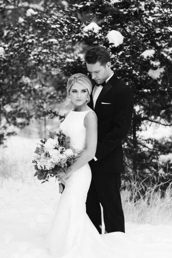 A bride in a white dress holding a bouquet stands in snowy outdoors, while a groom in a dark suit stands close behind her, both looking serene among snow-covered trees. by Justin Salem Meyer