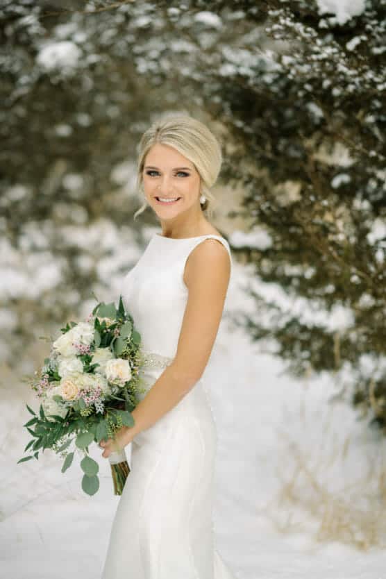 A bride in a sleeveless white wedding dress holds a bouquet of white and blush flowers, standing outdoors in a snowy, wooded area with trees in the background. She is smiling and looking at the camera. by Justin Salem Meyer