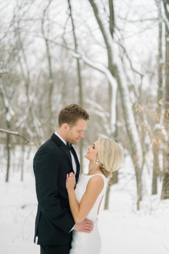 A bride and groom stand close together in a snowy forest, gazing into each others eyes. The groom wears a black tuxedo and the bride wears a white dress with buttons down the back. Snow covers the ground and trees behind them. by Justin Salem Meyer