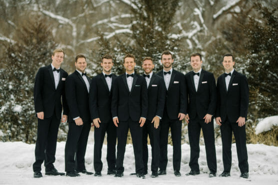Eight men in black tuxedos stand outside in the snow, smiling at the camera. Behind them are trees with bare branches and a wintery background. by Justin Salem Meyer