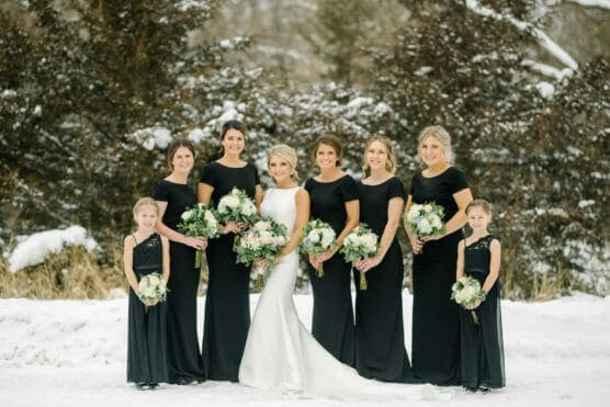 Eight women in black dresses, one in a white gown, stand outside on snow holding bouquets. They are posing for a photograph with trees in the background, suggesting a winter wedding scene. by Justin Salem Meyer