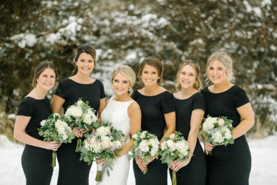 Six women stand side by side outdoors in winter, holding white and green bouquets. Five wear matching black dresses, and one in the center wears a white dress. Snow and trees fill the background. by Justin Salem Meyer
