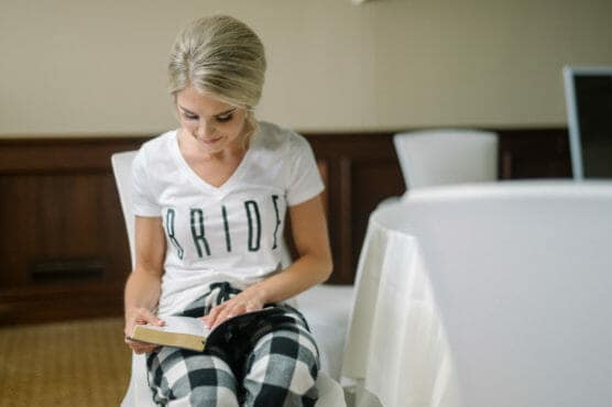 A woman with blonde hair in an updo, wearing a white BRIDE t-shirt and plaid pants, sits on a white chair and reads a small book at a round table in a softly lit room. by Justin Salem Meyer