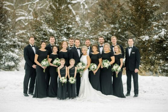 A wedding party poses outdoors in the snow. The bride, in a white dress, stands with bridesmaids in black dresses and groomsmen in black suits with bow ties. Everyone is holding bouquets, with snowy trees in the background. by Justin Salem Meyer