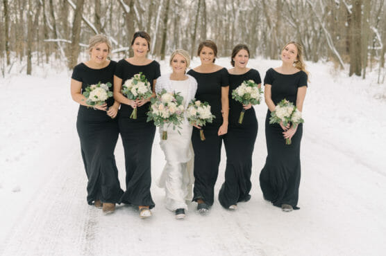 Six women stand together on a snowy path surrounded by trees. The bride, in a white gown, is flanked by five bridesmaids in matching black dresses, all holding bouquets and smiling. by Justin Salem Meyer