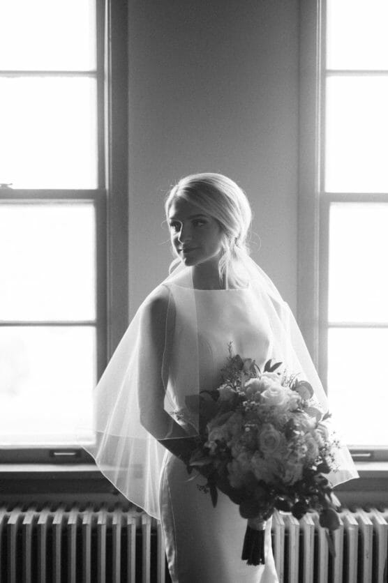 Black and white photo of a bride in a sleeveless gown and veil, standing indoors by tall windows, holding a bouquet of flowers and looking to the side. Soft natural light highlights her face and dress. by Justin Salem Meyer