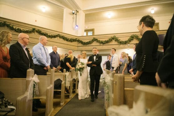 A bride in a white dress walks down the aisle, holding a bouquet and escorted by an older man, while guests in formal attire stand and watch inside a decorated church. by Justin Salem Meyer