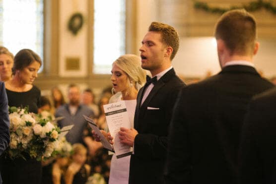 A man in a tuxedo and a woman in a white dress sing and hold programs at a wedding ceremony inside a church, surrounded by other well-dressed guests and floral arrangements. by Justin Salem Meyer