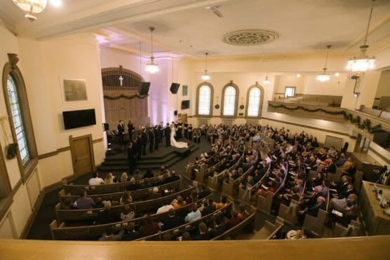 A wide view of a church interior during a wedding ceremony. Guests are seated in pews facing a couple at the altar, surrounded by attendants. Warm lighting streams in from arched windows, and greenery decorates the balcony railing. by Justin Salem Meyer