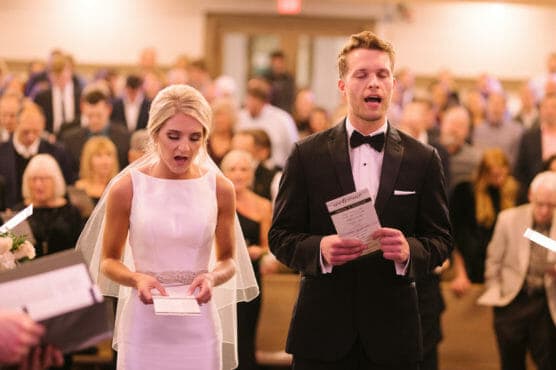 A bride and groom stand side by side in formal attire, holding programs and singing during a wedding ceremony with a crowd of seated and standing guests in the background. by Justin Salem Meyer
