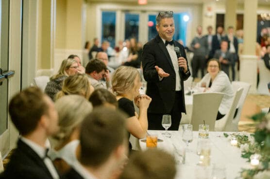A man in a tuxedo stands and speaks into a microphone at a formal event, addressing seated guests at candlelit tables in a banquet hall. People listen attentively and appear engaged. by Justin Salem Meyer