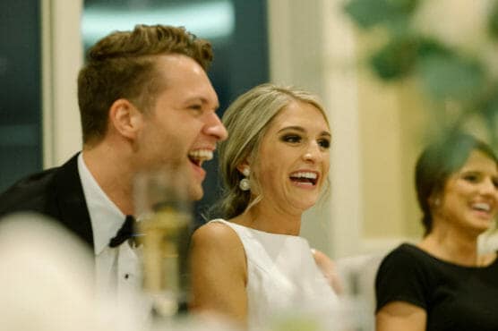 A man in a tuxedo and a woman in a white dress sit closely together, both smiling and laughing, with another woman in a black dress beside them. The atmosphere appears joyful and lively. by Justin Salem Meyer