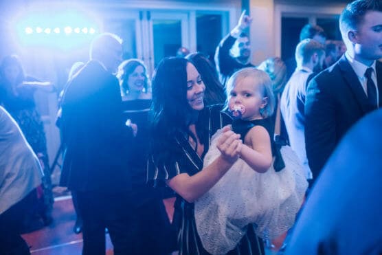 A woman in a striped dress smiles as she holds and dances with a young girl in a white dress and pacifier. Blue lighting and several people in formal wear fill the lively background. by Justin Salem Meyer
