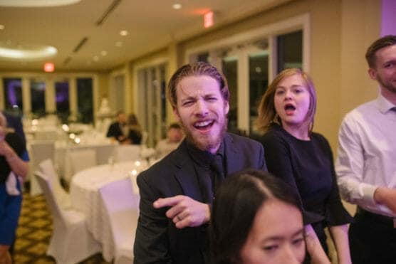 A bearded man in a dark suit laughs and points while standing next to a woman in a black dress at a lively indoor event with white table settings in the background. by Justin Salem Meyer
