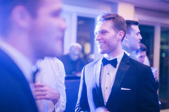 A man in a black tuxedo and bow tie smiles while talking with others at a formal indoor event, with blurred people and soft lighting in the background. by Justin Salem Meyer