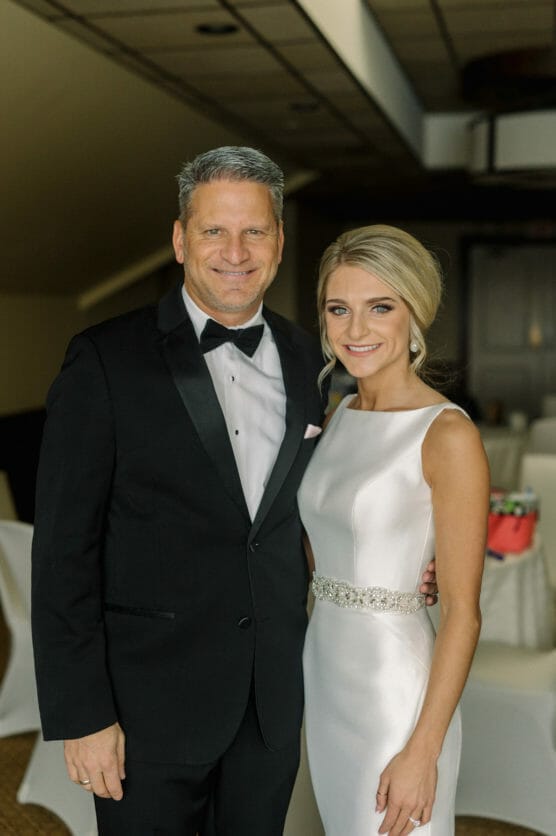A man in a black tuxedo stands next to a woman in a white sleeveless wedding dress. They are both smiling and posing indoors, possibly at a wedding reception. by Justin Salem Meyer