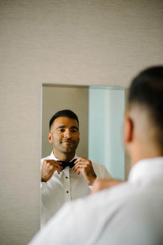 A man in a white dress shirt adjusts his black bow tie while looking at himself in a mirror, smiling slightly. The photo captures his reflection as he prepares, with a neutral background behind him. by Justin Salem Meyer