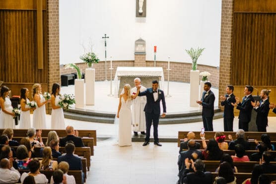 A bride and groom stand at the altar of a church, holding hands and smiling. Bridesmaids in white dresses and groomsmen in dark suits stand on either side, while guests seated in pews look on and applaud. by Justin Salem Meyer
