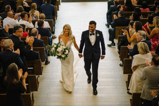 A bride in a white dress and a groom in a dark suit walk down the aisle holding hands, smiling, as wedding guests seated on both sides clap and celebrate their marriage in a bright indoor venue. by Justin Salem Meyer