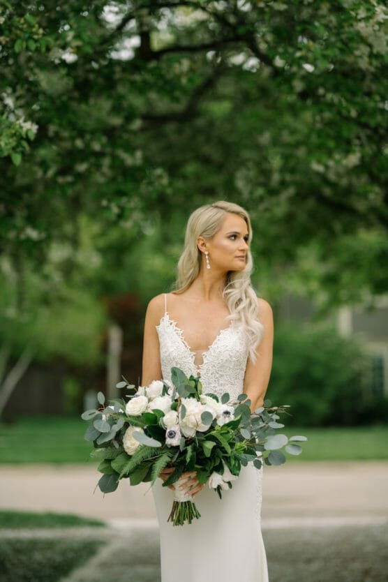 A bride in a white gown with lace details holds a large bouquet of white flowers and greenery, standing outdoors with lush trees in the background. She has long blonde hair and looks to the side. by Justin Salem Meyer