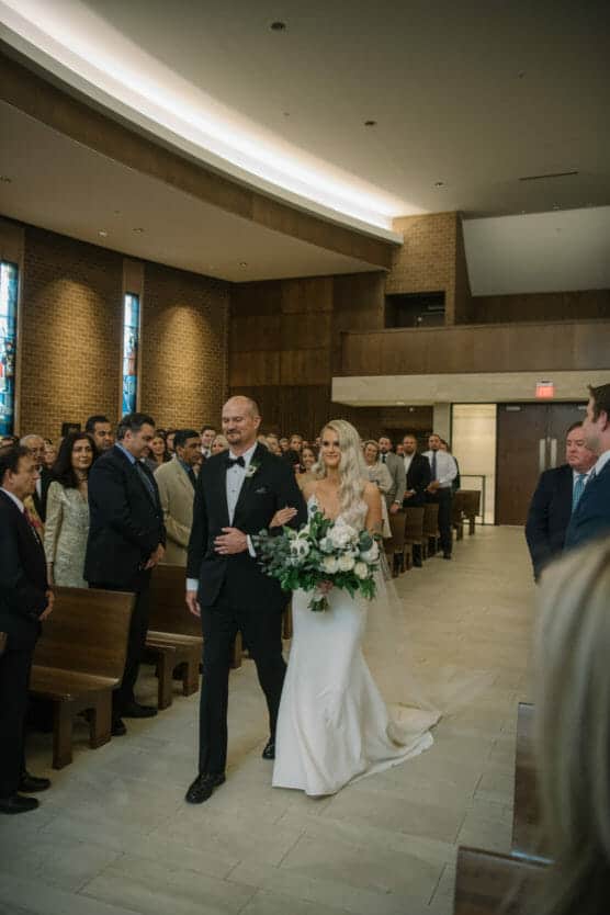 A bride in a white dress holding a bouquet walks down the aisle with a man in a tuxedo, surrounded by guests seated in wooden pews in a church. by Justin Salem Meyer