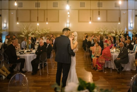 A bride and groom share their first dance in the center of a warmly-lit reception hall, surrounded by seated guests at round tables decorated with white flowers and candles. by Justin Salem Meyer