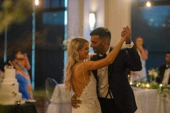 A bride and groom share a joyful first dance at their wedding reception, holding hands and smiling at each other. The bride wears a white dress with lace details, and the groom is in a dark tuxedo. The room is softly lit. by Justin Salem Meyer