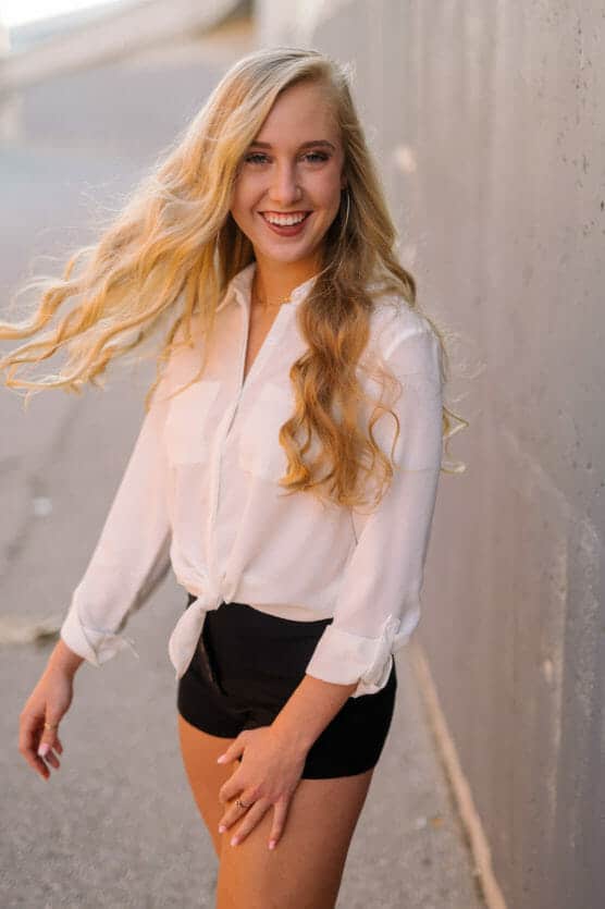 A young woman with long, wavy blonde hair smiles brightly while standing outdoors. She wears a white blouse tied at the waist and black shorts, with one hand slightly raised and loose hair flowing around her. by Justin Salem Meyer