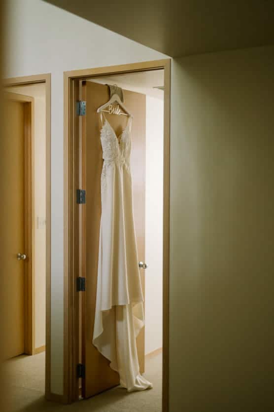 A white bridal gown with lace detailing hangs on a wooden hanger labeled Bride, displayed on a door in a softly lit hallway with beige walls and carpet. by Justin Salem Meyer