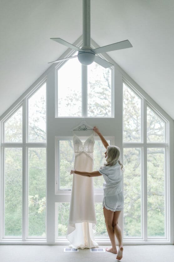 A woman in pajamas stands barefoot in a bright room, reaching up to a wedding dress hanging from a hanger labeled Bride in front of large windows with a green, leafy view outside. by Justin Salem Meyer