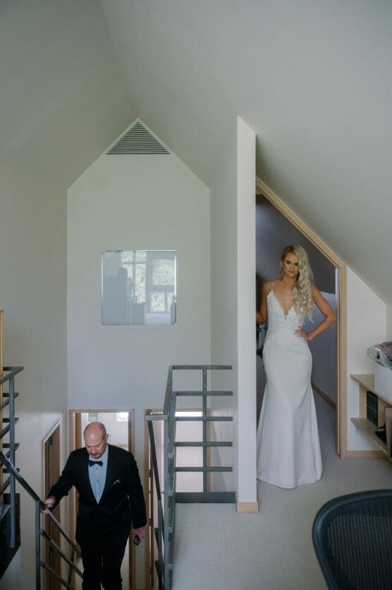 A bride in a white gown stands in a doorway at the top of a staircase, while a man in a tuxedo ascends the stairs in a modern, minimalist interior with slanted ceilings. by Justin Salem Meyer