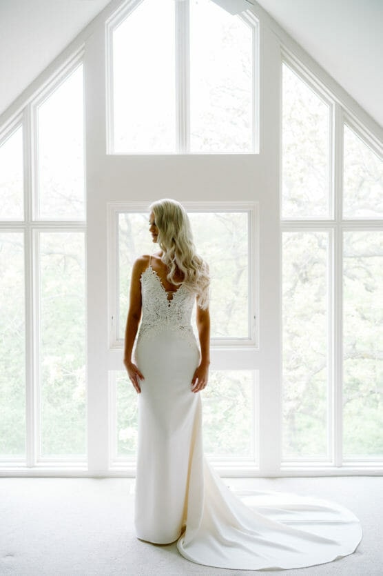 A bride with long, wavy blonde hair stands in front of large, bright windows wearing a white, fitted wedding dress with a lace back and train, looking away from the camera. by Justin Salem Meyer