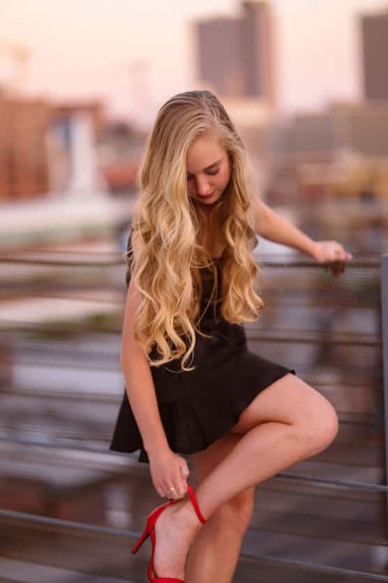 A young woman with long, wavy blonde hair in a black dress sits on a railing, looking down while adjusting her red high-heeled shoe. The cityscape in the background is softly blurred. by Justin Salem Meyer