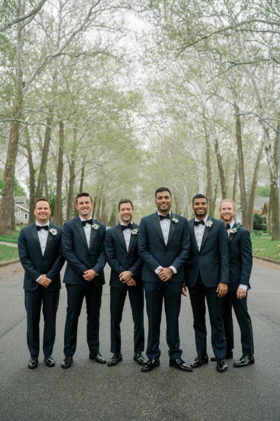 Six men wearing matching dark suits with bow ties stand side by side on a tree-lined street, smiling at the camera. The background features tall, leafless trees and overcast skies. by Justin Salem Meyer