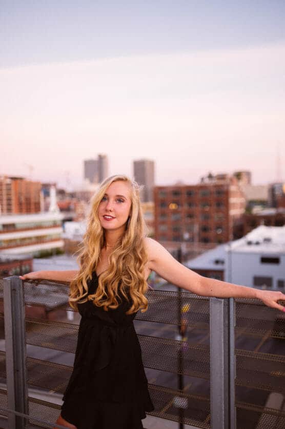 A young woman with long blonde hair, wearing a black dress, stands on a rooftop at sunset with city buildings in the background, resting her arms on a metal railing and looking at the camera. by Justin Salem Meyer