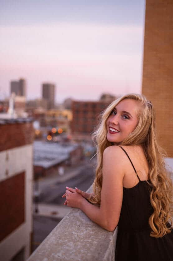 A young woman with long blonde hair in a black dress leans on a rooftop ledge, smiling toward the camera. The city skyline and soft pink sunset are visible in the background. by Justin Salem Meyer