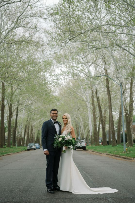 A bride in a white dress and a groom in a dark suit stand together smiling on a tree-lined street, holding a bouquet of greenery, with parked cars and leafing trees in the background. by Justin Salem Meyer