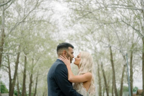 A couple embraces outdoors beneath tall, leafy trees. The woman, with long blonde hair in a white dress, gently holds the mans face, while he wears a dark suit. They gaze lovingly into each others eyes. by Justin Salem Meyer