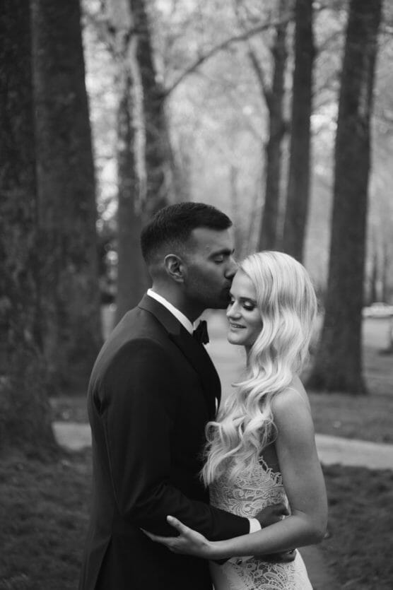A groom in a suit gently kisses the forehead of his smiling bride in a lace dress as they embrace outdoors among tall trees. The image is in black and white. by Justin Salem Meyer