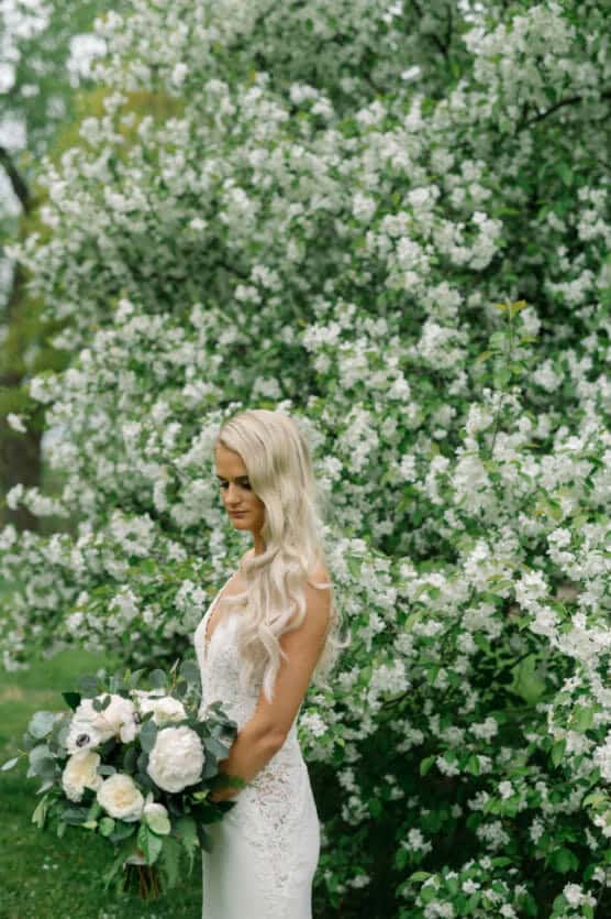 A bride in a white lace dress with long, wavy blonde hair holds a bouquet of white and green flowers, standing in front of a lush blooming tree covered in small white blossoms. by Justin Salem Meyer