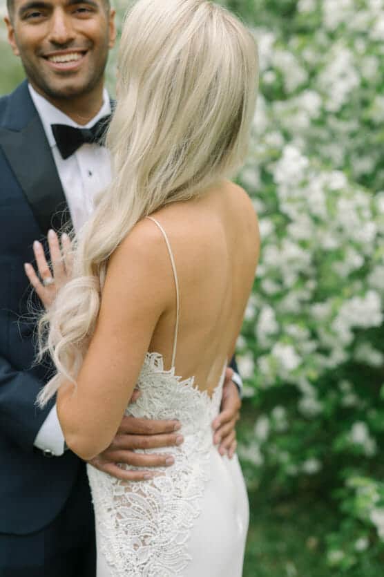 A bride in a white lace dress with long, wavy blonde hair embraces a groom in a dark suit and bow tie, both smiling, in front of a backdrop of lush white flowers and greenery. by Justin Salem Meyer