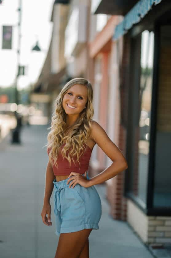 A young woman with long, wavy blonde hair, wearing a sleeveless maroon top and light blue high-waisted shorts, smiles while standing on a sunny sidewalk with storefronts in the background. by Justin Salem Meyer